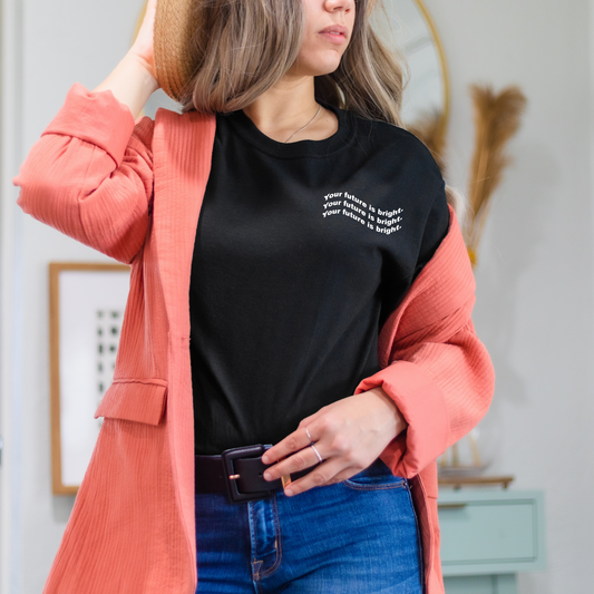 Girl with light brown hair looking off into the distance wearing a hat, coral open jacket, jeans, and a black t-shirt with white writing that says "your future is bright".